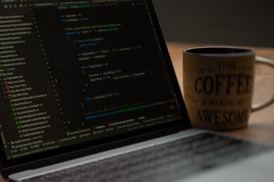 A close-up view of a person's hand with a wristwatch, typing on a silver laptop placed on a wooden desk. The laptop screen displays lines of code with a dark background and colored syntax highlighting